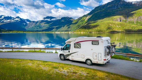 A RV with bikes attached driving next to a lake with beautiful mountains in the background.