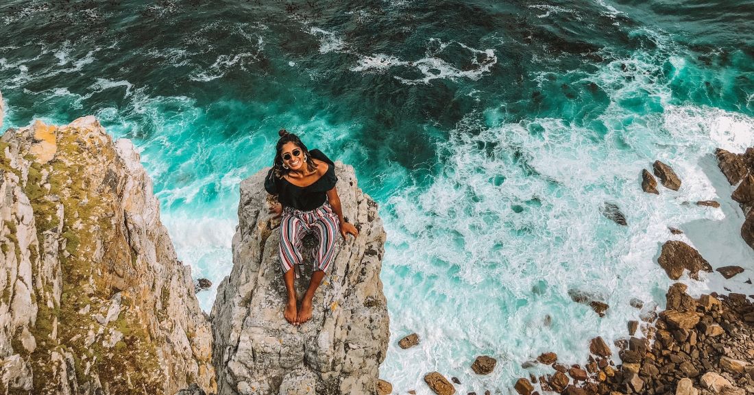 Dr Nabila Ismail sitting on a rocky overhang with the ocean waves crashing below -- a presentation for the Wanderful community on traveling solo on a budget